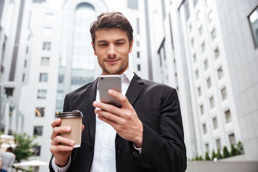 Handsome, well-dressed young man in urban setting, holding his phone and a coffee to go.